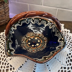 May include: A decorative ceramic bowl with a dark blue interior and a brown, twisted handle. The bowl features a detailed, colorful floral pattern in the center and along the rim, with white, green, and orange accents. The rim has a wavy design.