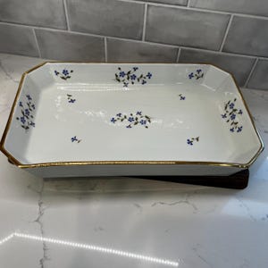 May include: A rectangular white ceramic serving tray with beveled corners and a gold-toned rim. The interior surface features a delicate pattern of small blue flowers with green stems and leaves scattered across the base and along the sides.