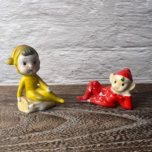 May include: Two ceramic figurines of elves. One elf is in a yellow outfit and sitting on a beige base. The other elf is in a red outfit and is lying down. Both have white faces and are painted with details.