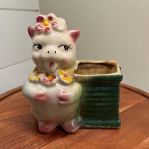 May include: A ceramic planter shaped like a pig wearing a flower crown. The pig is white with pink accents and has a green planter attached to its back. The planter has a brown interior and is ready for planting.