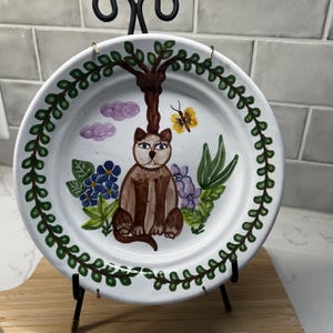 May include: A white ceramic plate with a hand-painted cat design. The cat is brown with blue eyes, sitting under a tree with a yellow butterfly. The plate has a green leaf border and is displayed on a black metal stand.