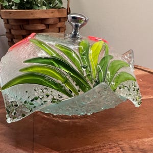 May include: A clear glass serving dish with a silver knob on the lid. The dish is decorated with green and pink glass leaves.