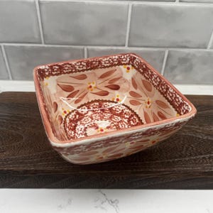 May include: Square ceramic bowl with a floral design in shades of peach, brown, and cream. The bowl has a decorative rim and is set on a dark wooden surface. The interior features a detailed, symmetrical pattern.