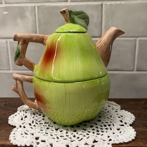 May include: A ceramic tea set for one, shaped like a pear with a light green and red finish. The top section is a teapot with a brown branch-like handle and spout, while the bottom section is a matching teacup with a branch-shaped handle.