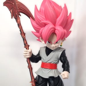 Op de afbeelding: Een verzamelbaar anime-figuur met een personage met stekelig roze haar, gekleed in een zwart shirt met lange mouwen, een grijs vest, een rode sjerp en een zwarte broek. Het figuur houdt een grote, metallic rode zeis vast.