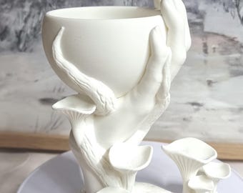 May include: A white ceramic sculpture of a hand holding a bowl, with mushroom-like structures at the base. The sculpture is on a white plate, set against a blurred backdrop of a winter landscape. The artwork is a unique decorative piece.