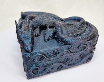 May include: A black heart-shaped box with a dragon design. The dragon is curled up on the lid of the box, with its wings spread out. The box is made of a textured material and has a detailed design.