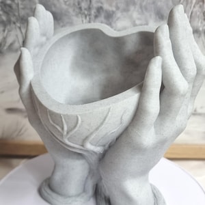 Puede incluir: Una maceta gris en forma de manos con una abertura en forma de corazón. La maceta está hecha de un material moteado y descansa sobre una superficie blanca. El fondo presenta una imagen borrosa de un paisaje invernal.