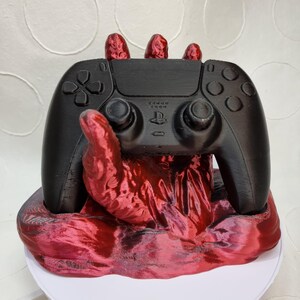 May include: A black PlayStation 5 controller is held by a 3D printed red hand. The hand is resting on a red base.