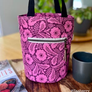 May include: A cylindrical tote bag with a floral pattern in shades of pink and purple. The bag has black handles and a zippered front pocket with a white zipper. A small leather tassel hangs from the zipper pull.