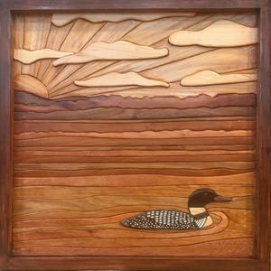 Könnte beinhalten: Ein quadratisches Wandkunstwerk aus Holz mit einer Intarsien-Landschaftsszene. Das Bild zeigt einen Eistaucher, der auf dem Wasser schwimmt, mit einem Sonnenaufgang und Wolken im Hintergrund. Das Kunstwerk besteht aus verschiedenen hellen und dunklen Holztönen.