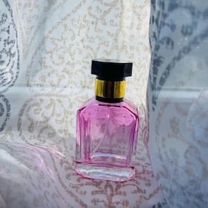 May include: A pink glass perfume bottle with a black and gold cap. The bottle is sitting on a white fabric background.