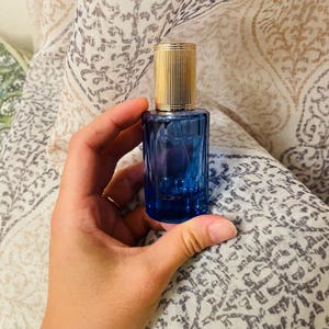 Può includere: Un flacone di vetro blu con un tappo dorato e un liquido trasparente all'interno. Il flacone è tenuto in una mano.