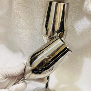May include: Two polished silver champagne flutes with a slender stem and a wide bowl. Each flute has an engraved cursive design. The flutes are displayed on a white surface.