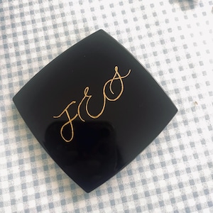 May include: A black, square compact with rounded corners. The compact has gold cursive lettering that reads "JRS". The compact is on a light blue and white checkered surface.