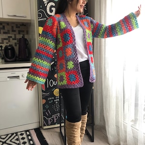 Puede incluir: Un cárdigan de crochet colorido con un diseño de patchwork. El cárdigan tiene mangas largas y un corte holgado. La persona que lleva el cárdigan está de pie en una cocina.