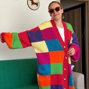 以下が含まれることがあります： 黄色、赤、オレンジ、緑、青、紫の大きな四角形を組み合わせた鮮やかなパッチワーク柄の厚手のニットカーディガン。長袖でVネックのデザインとなっており、前面にはカラフルな丸いボタンが並び、ゆったりとしたフィット感のあるシルエットです。