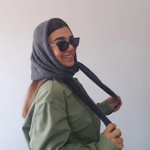 Puede incluir: Un pañuelo de punto gris oscuro atado bajo la barbilla, combinado con gafas de sol negras estilo ojo de gato y una camisa verde oliva con botones, dos bolsillos en el pecho y mangas largas. El pañuelo tiene una apariencia texturizada.