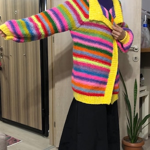 May include: A long, open-front cardigan with horizontal rainbow stripes. The collar and trim are bright yellow. The sleeves are long and the sweater appears to be handmade. The garment is worn over a black skirt.