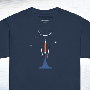 May include: Navy blue t-shirt featuring a minimalist graphic of a rocket launching towards a crescent moon and stars. The design includes orange and white elements, with the text "DIGITAL ASTRONAUT" on the tag.