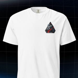 Puede incluir: Camiseta blanca con un pequeño parche triangular en el pecho. El parche presenta un diseño con elementos grises y rojos, y el texto "ROCKET LAUNCHERS". La camiseta tiene mangas cortas y cuello redondo.