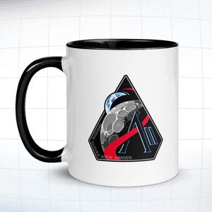 May include: White ceramic mug with a black handle and interior. The mug features a triangular design with a depiction of the Earth, the moon, and the letter "A". The names "WISEMAN", "GLOVER", "KOCH", and "HANSEN" are printed on the design.