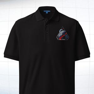 May include: Black polo shirt with a small embroidered patch on the left chest. The patch features a geometric design with a space-themed illustration in shades of blue, red, and white. The shirt has a classic collar and buttoned placket.