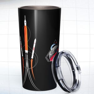 May include: Black travel tumbler with a rocket ship design. The tumbler features a black lid and a graphic of a rocket ship with orange and white accents. The NASA logo is visible on the side of the rocket. The tumbler is cylindrical.