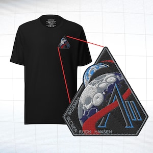 May include: Black t-shirt with a small embroidered patch featuring a space-themed design. The patch includes a stylized moon, Earth, and the names WISEMAN, GLOVER, and KOCH HANSEN. The design incorporates red, blue, and white colors.