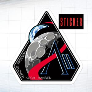 May include: A sticker featuring a black and white graphic of the moon, Earth, and a red and blue design. The sticker has the word "STICKER" in red and the names "WISEMAN", "GLOVER", "KOCH", and "HANSEN".