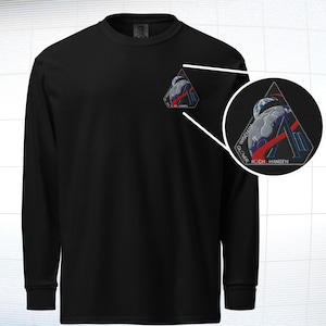 May include: Black long-sleeve shirt with a small embroidered patch on the chest. The patch features a triangular design with a stylized image of a planet and the names "WISEMAN, GLOVER, KOCH, HANSEN". The shirt has a crew neck and ribbed cuffs.