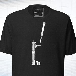 May include: Black t-shirt with a white graphic design featuring a syringe and a structure resembling an airport control tower. The shirt has a tag in the upper center with text. The design is a minimalist, modern style.