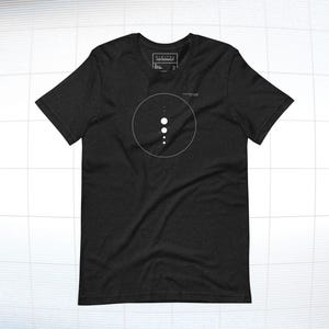 Puede incluir: Camiseta gris oscuro con un diseño circular blanco y puntos blancos. La camiseta tiene cuello redondo y mangas cortas. El nombre de la marca está impreso en blanco en la esquina superior izquierda.