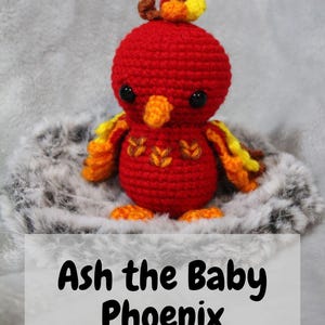 Könnte beinhalten: Ein handgehäkeltes rotes Baby-Phönix mit gelben und orangen Akzenten. Das Amigurumi hat schwarze Augen und einen kleinen Schnabel. Der Text "Ash the Baby Phoenix" wird darunter angezeigt, mit "PDF AMIGURUMI PATTERN".