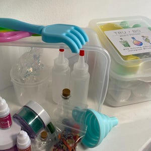 May include: A clear plastic container filled with various craft supplies. Includes bottles, jars, glitter, a blue funnel, and a hand-shaped scoop. A box labeled "TRU + BO MAGIC POTION" is also visible.