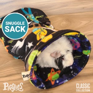 Può includere: Un Snuggle Sack nero e colorato con un motivo spaziale, con pianeti, stelle e astronauti. Un piccolo porcellino d'India bianco e nero è all'interno. Il testo "Snuggle Sack" è in un cerchio blu.