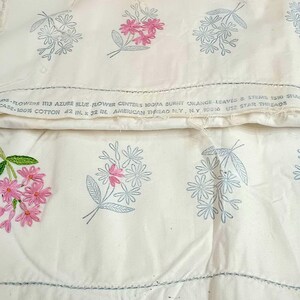 May include: Two off-white cotton pillowcases featuring embroidered floral motifs in shades of pink and light blue. The fabric includes printed text stating "AMERICAN THREAD N.Y. 10018" and "42 IN. X 36 IN.", indicating the dimensions of the pillowcases.