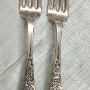 May include: Two antique silver forks with four tines each. The handles feature ornate floral and geometric designs, with an initial "ME" at the base. The forks are displayed on a light blue cloth.