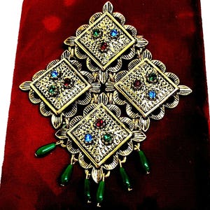 May include: Ornate gold-toned brooch with four diamond-shaped sections, each adorned with colorful gemstones. Green teardrop-shaped beads dangle from the bottom. The brooch rests on a deep red velvet surface.