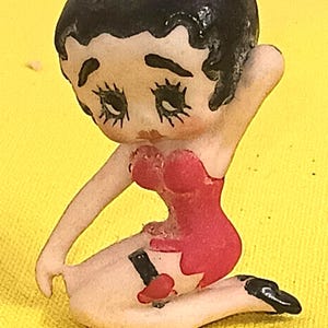 May include: A vintage figurine of a cartoon woman with black hair and a red dress. The figurine is kneeling with one leg bent and the other extended. The figurine has black painted shoes and is set against a yellow background.