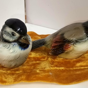 May include: Two ceramic bird figurines, one with a black cap and the other with a gray and brown body, rest on a gold velvet base. The birds have detailed features and are positioned side-by-side, showcasing their delicate craftsmanship.