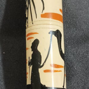 May include: A cylindrical container with a cream-colored background, featuring black silhouettes of a palm tree, a camel, and a person with a staff. Orange accents are present. The container is likely for storage or display.