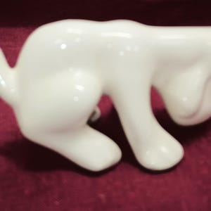 May include: A white ceramic figurine of a dog in a playful pose. The dog is standing on all fours with its head tilted down and its tail curled up.