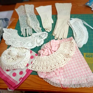 May include: A collection of vintage-style accessories including white lace gloves, solid white gloves, a white crocheted collar, a white crocheted hat, a white crocheted semi-circle piece, a pink and white polka dot scarf, and a pink gingham apron.