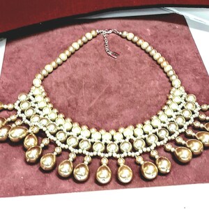May include: A gold and cream pearl necklace with a cascading design. The necklace is made of multiple strands of pearls, with the bottom strand featuring larger, teardrop-shaped pearls.