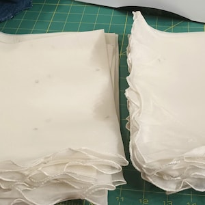 May include: Two stacks of off-white, square fabric pieces with ruffled edges. The fabric appears to be sheer, with a few small, dark specks visible. The pieces are neatly stacked, suggesting they are ready for use in a sewing or craft project.