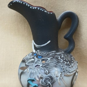 May include: A small black ceramic pitcher with a white dragon design and blue accents. The pitcher has a handle and a spout.