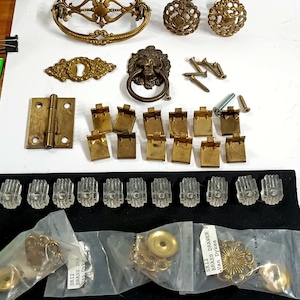 May include: A collection of vintage brass hardware components, including drawer pulls, knobs, hinges, and keyhole covers. The hardware features ornate designs and includes screws for installation. Some items are packaged in clear plastic bags.