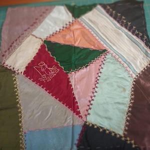 May include: A square patchwork textile piece featuring various fabric scraps in shades of red, green, pink, blue, and brown. The pieces are joined by decorative cross-stitching. A small embroidered figure of a person fishing is on a red patch.