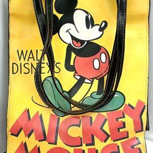 May include: Yellow tote bag featuring Mickey Mouse with black handles. The bag has the words "Walt Disney's" and "Mickey Mouse" in red lettering. Mickey Mouse is wearing red shorts, green shoes, and white gloves.